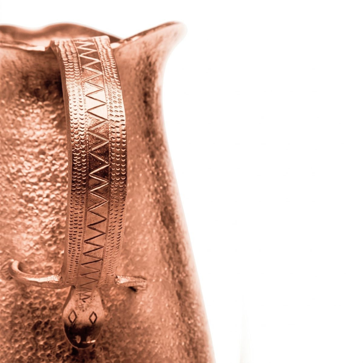 Close-up of a hammered copper pitcher with a decorative geometric handle, shown against a solid white background.