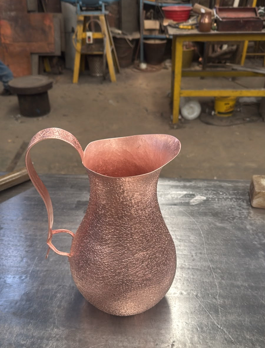 A hammered copper pitcher with a curved handle, sitting on a scratched metal workbench in a workshop environment.