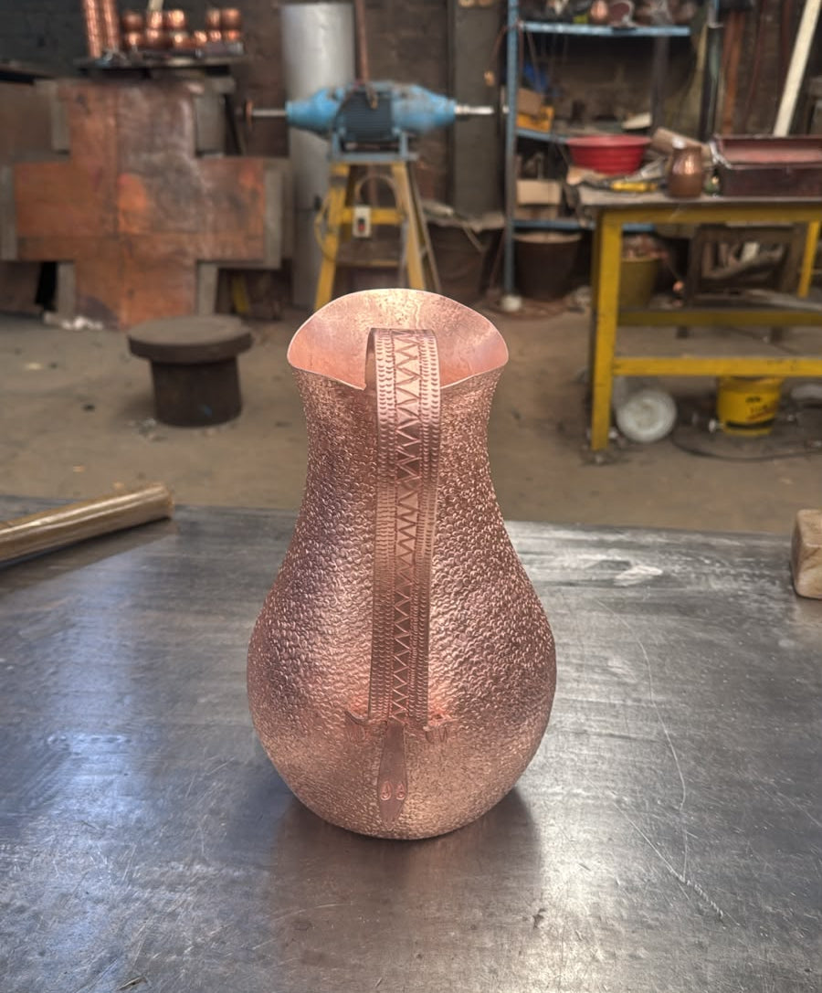 A hammered copper pitcher with a vertical handle, standing on a metal workbench in a workshop environment.