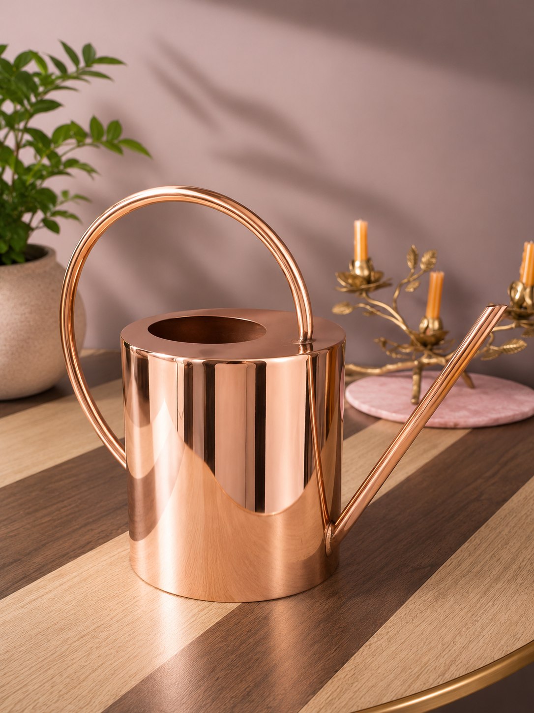 Copper watering can on a wooden surface with a plant and candle holder in the background.