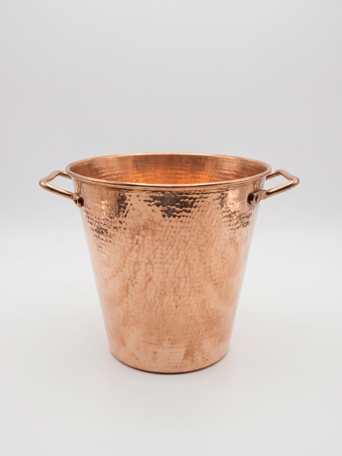 A hammered copper bucket with two side handles, photographed on a clean light gray studio background.