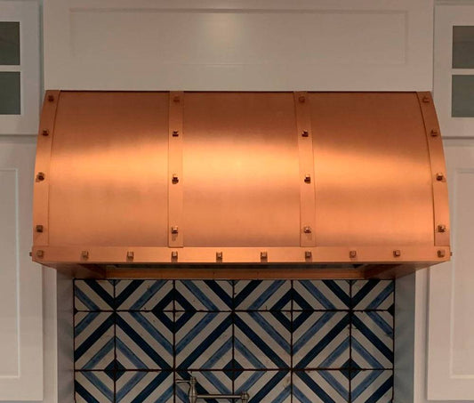 Amoretti Brothers WINSTON copper range hood, handcrafted solid copper with rivets, above geometric tile backsplash.